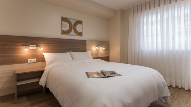 Una cama bien tendida con sábanas blancas, una mesita de noche con una lámpara y una revista sobre la cama.