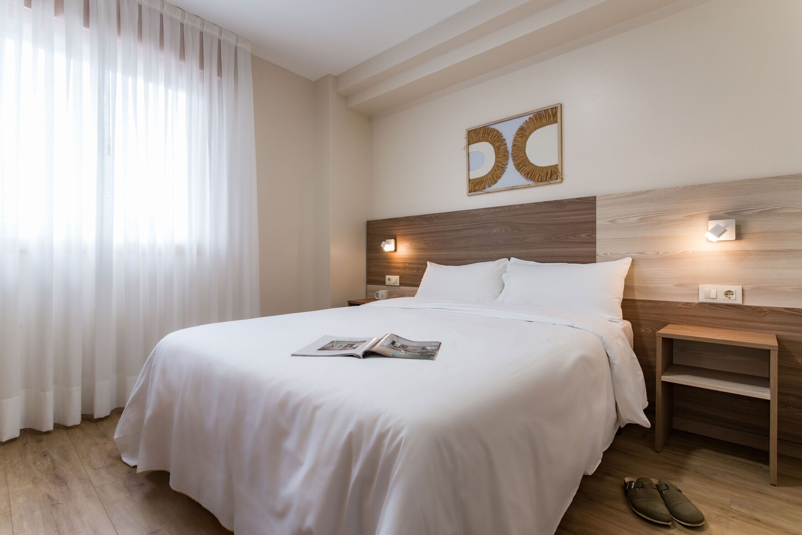 Una cama bien hecha con una revista, una mesita de noche de madera con una lámpara y un par de zapatos en el suelo.
