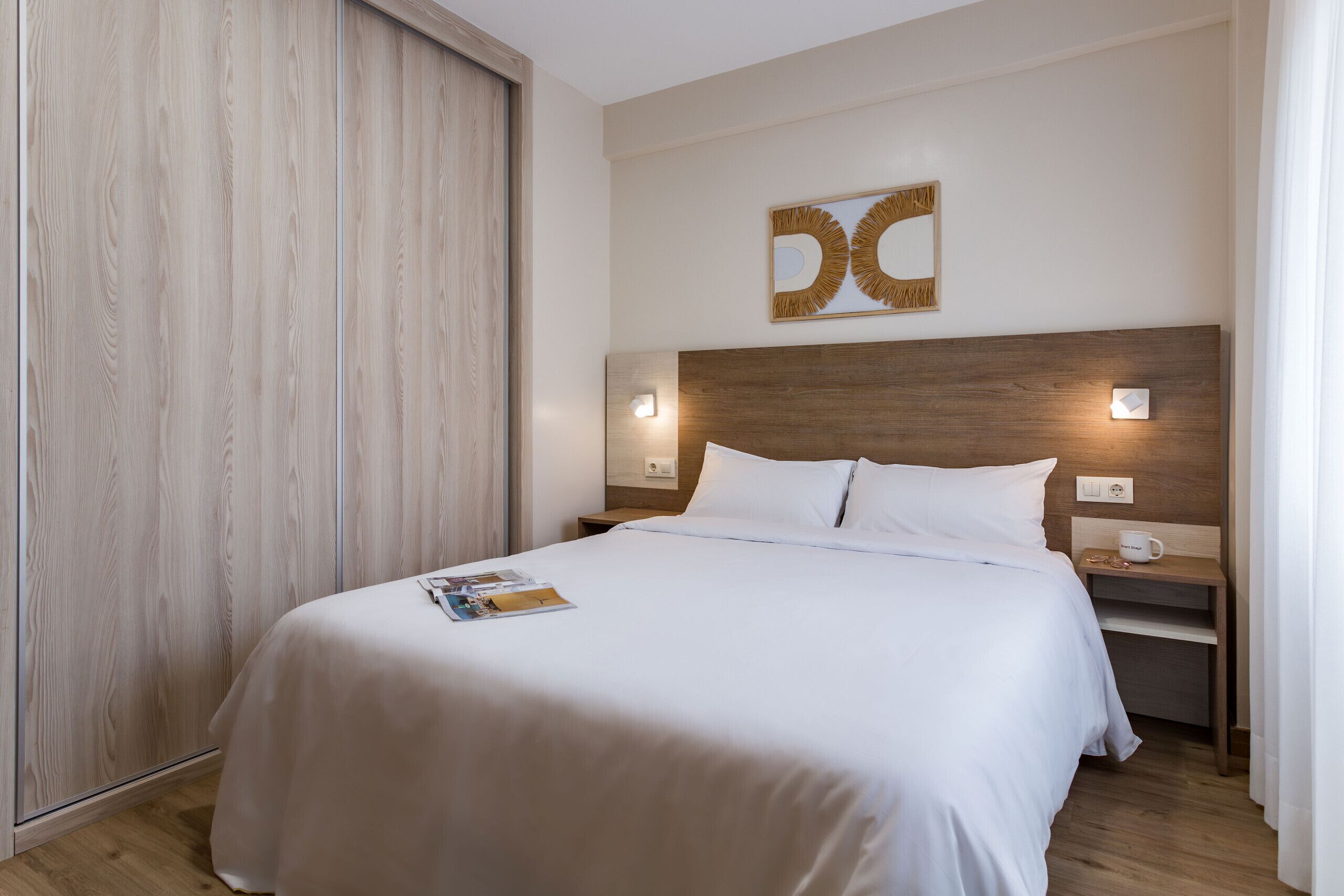 A hotel room with a wooden headboard, a bed with white linens, a magazine on the bed, a nightstand with a cup and a phone, and a wall-mounted light fixture.