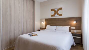 A hotel room with a wooden headboard, a bed with white linens, a magazine on the bed, a nightstand with a cup and a phone, and a wall-mounted light fixture.