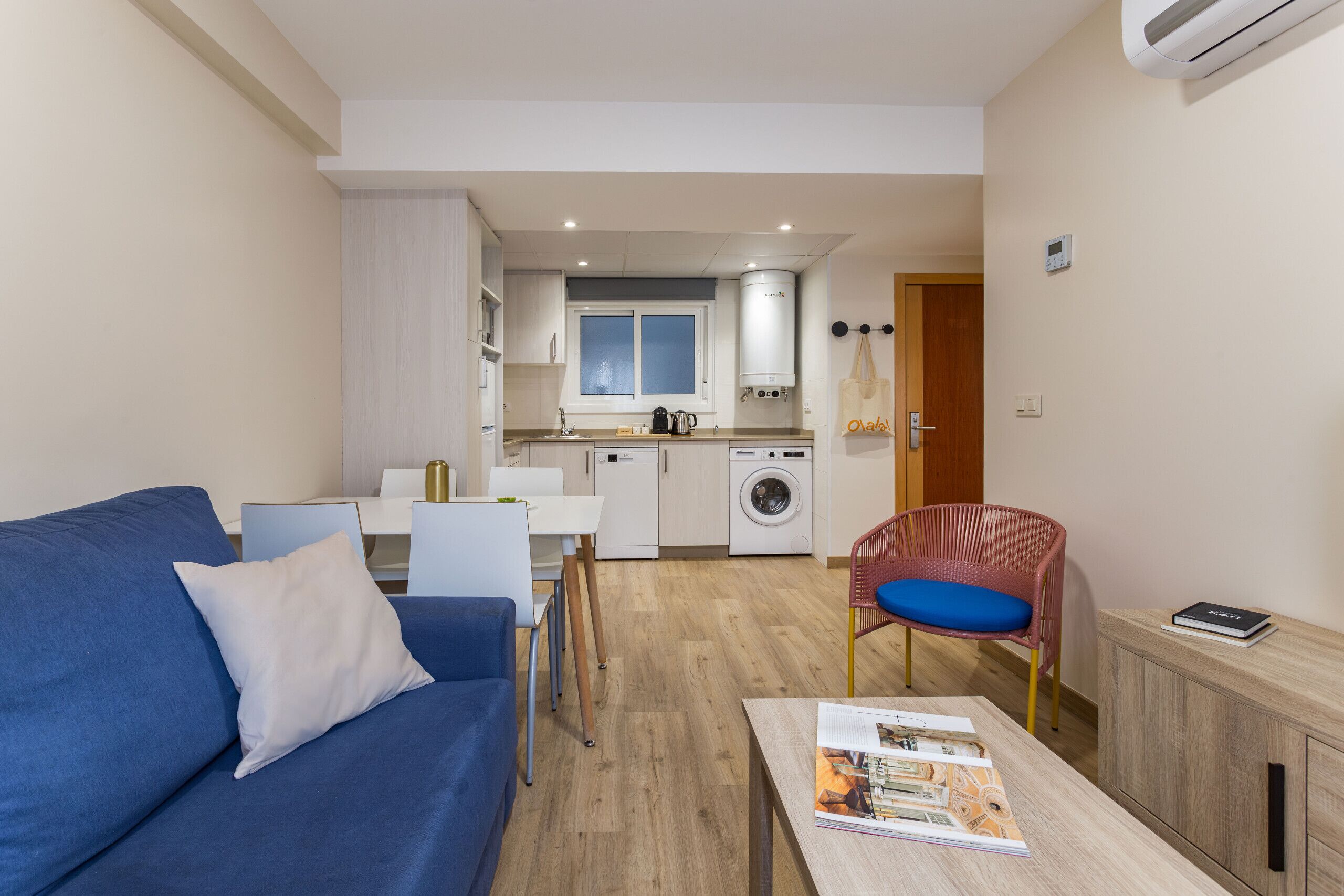 A compact living space with a blue sofa, a dining table, a kitchen area with a washing machine, and a small seating area with a chair.