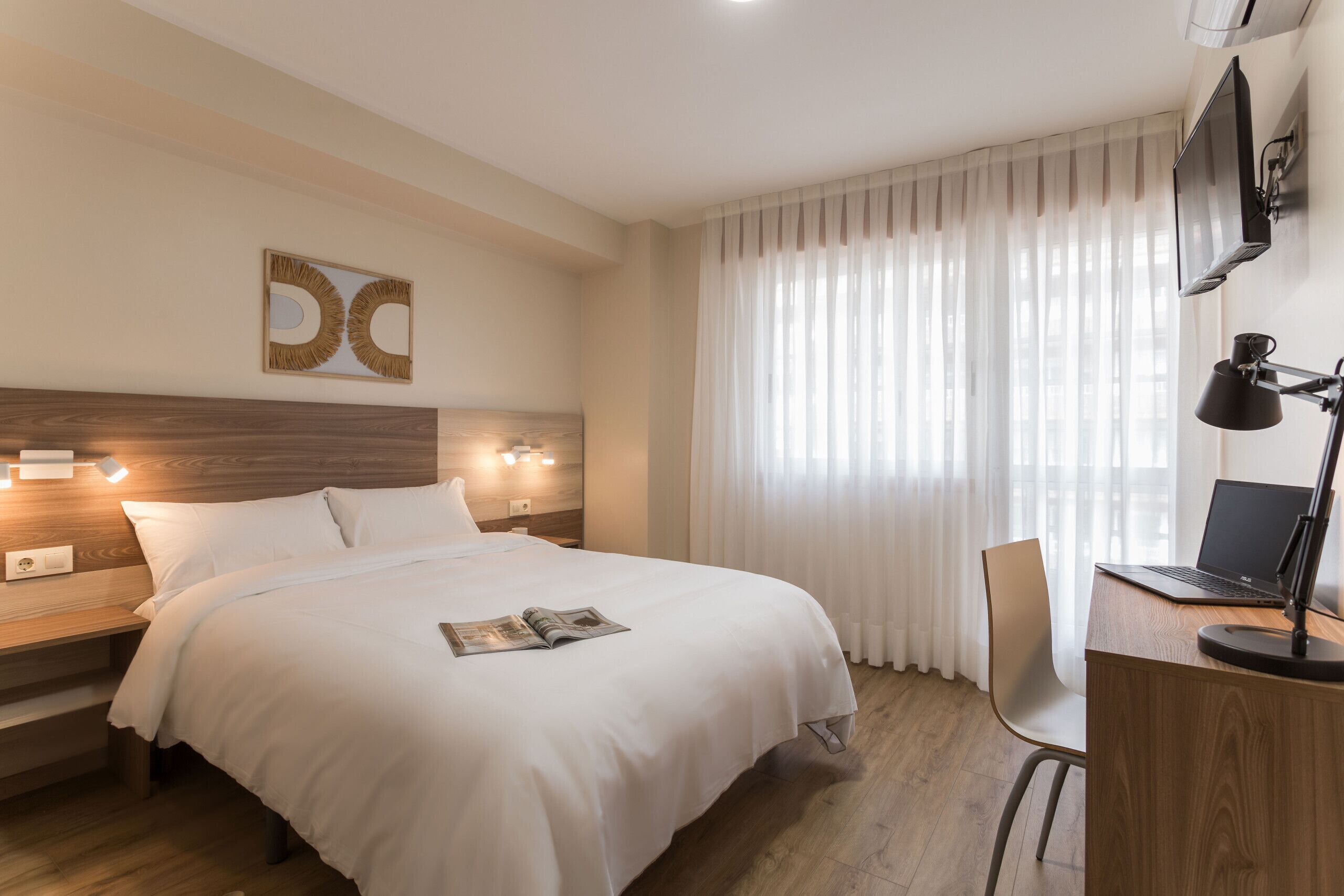 A hotel room with a bed, bedside tables, a desk with a laptop, a chair, and a TV.