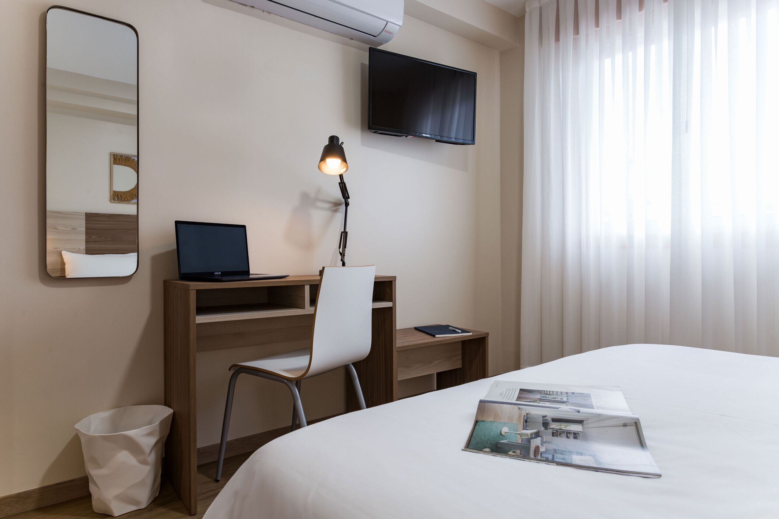 A hotel room with a bed, a desk with a laptop, a chair, a TV mounted on the wall, and a window with curtains.