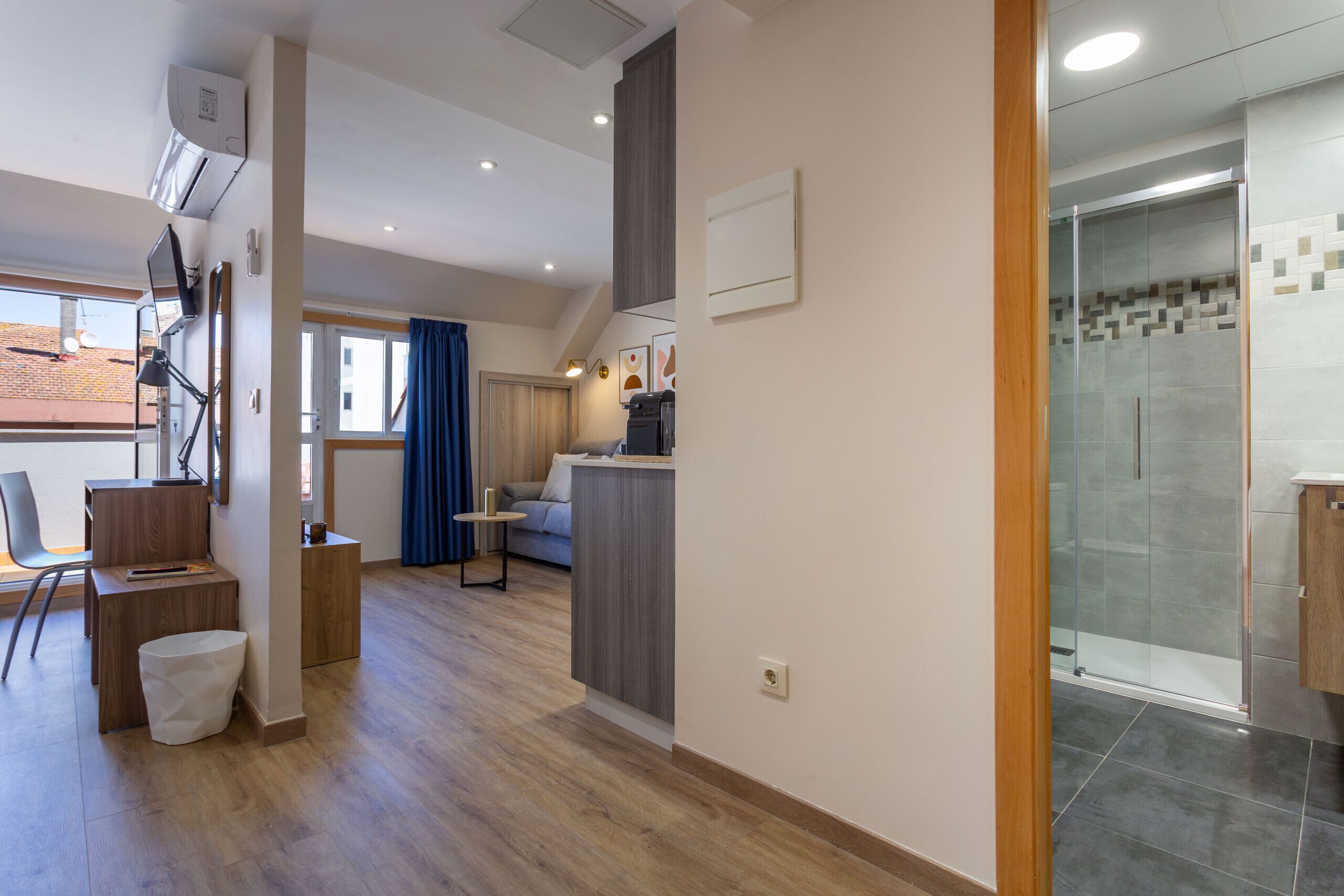 Una moderna camera d'hotel con una scrivania in legno, un piccolo angolo salotto, una doccia con pareti in vetro e vista esterna.
