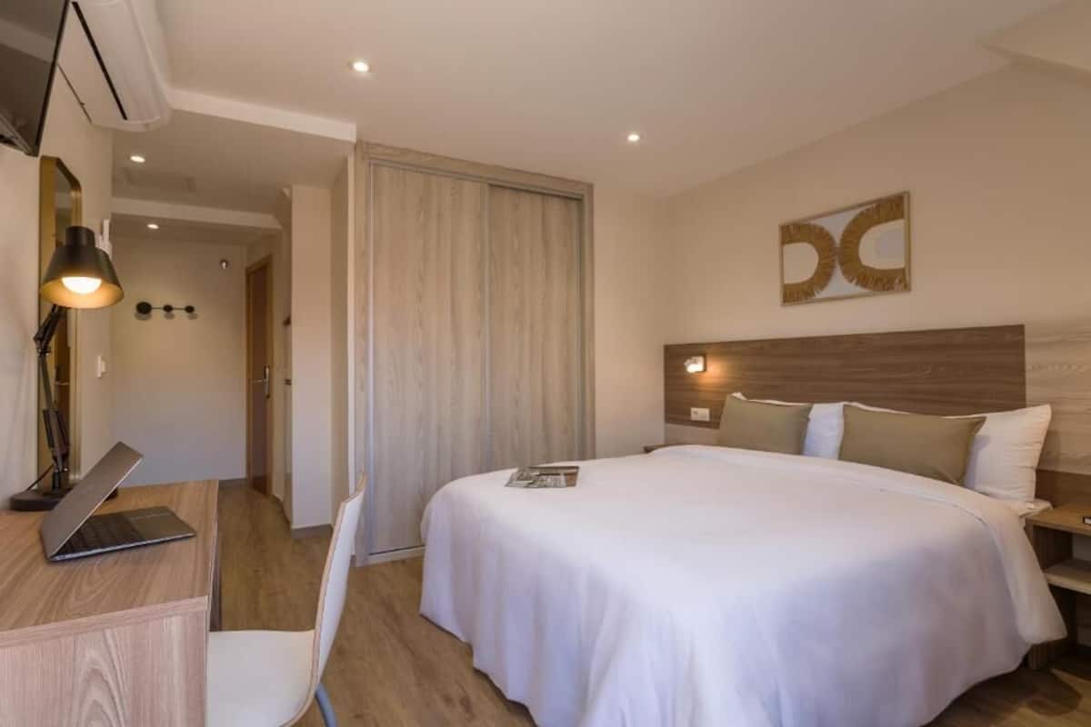 A hotel room with a bed, a desk with a laptop, a chair, and a door leading to another room.