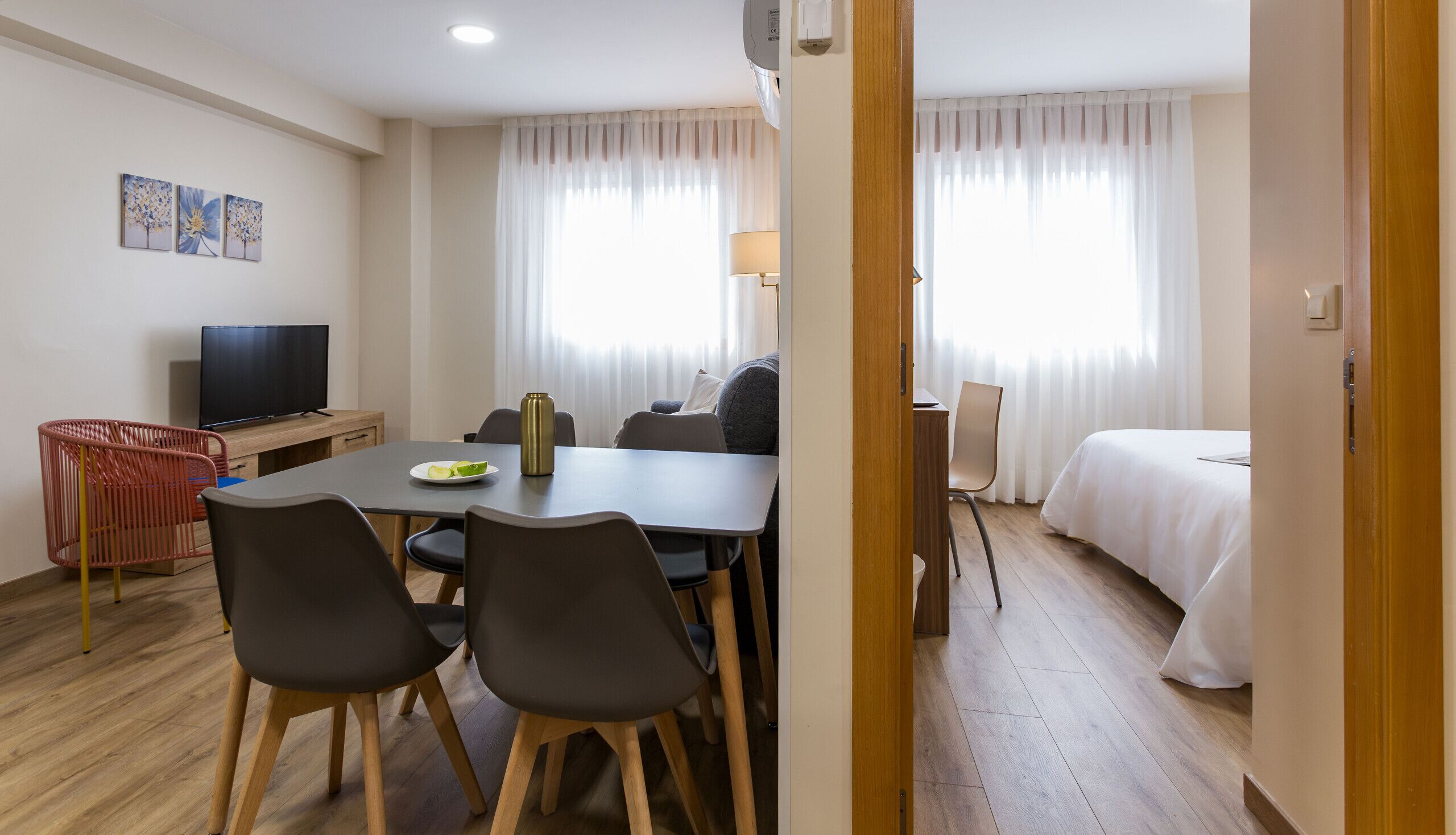 Habitación de hotel con cama, escritorio y silla, mesa de comedor con sillas, televisor y ventana con cortinas.