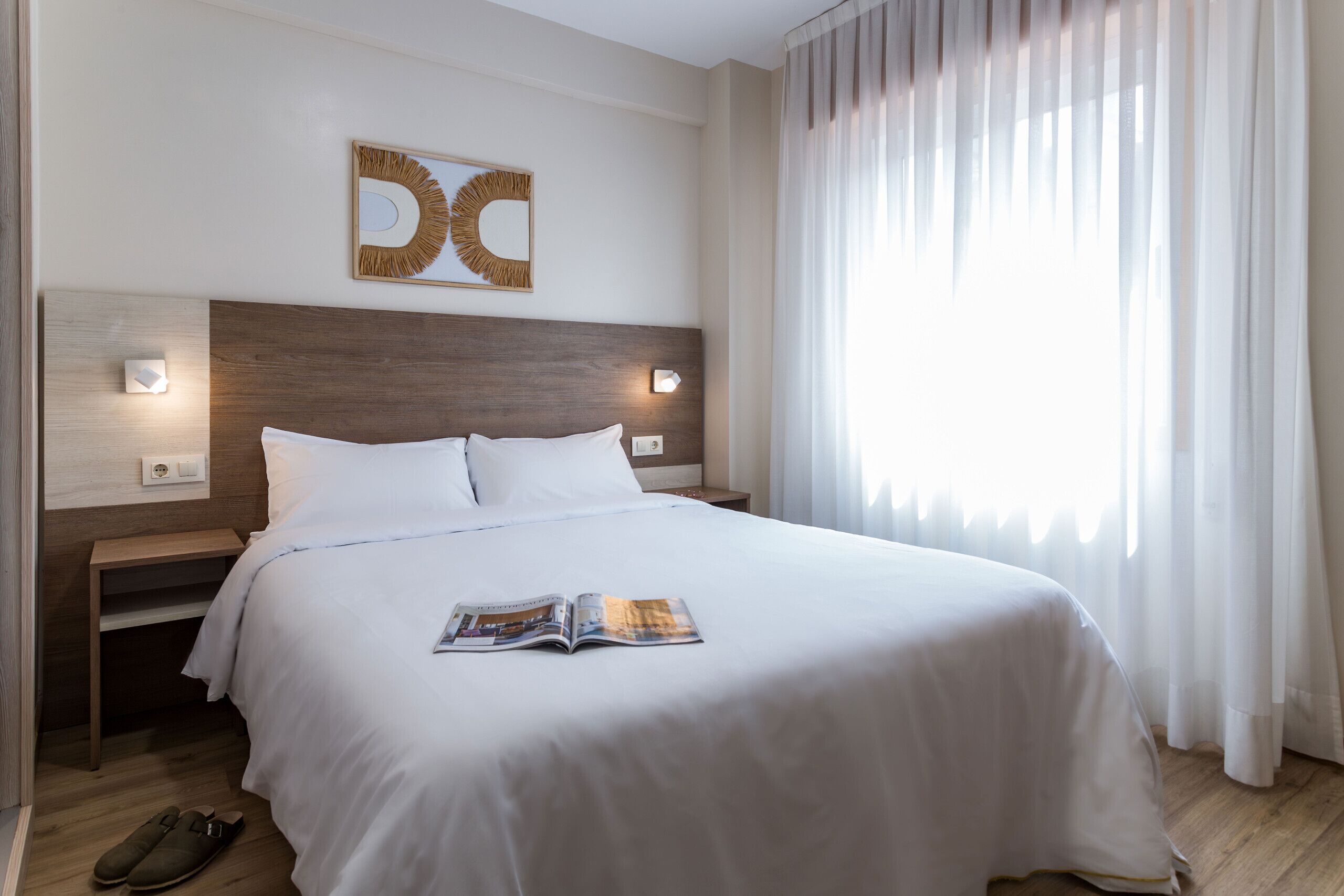 Una cama bien tendida con una revista encima, un cabecero de madera, dos lámparas de noche y un ventanal con cortinas ligeras.