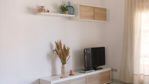 A modern living room with a green sofa, a dining table with a woven placemat, a TV on a white cabinet, and a potted plant on a shelf. - Tranquilidad, Naturaleza y Mar. Tu Refugio (L'Eucaliptus)