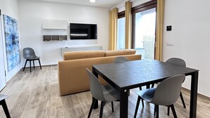 Una sala de estar moderna con un sofá, una mesa de comedor con sillas, un televisor montado en la pared y un ventanal con cortinas.