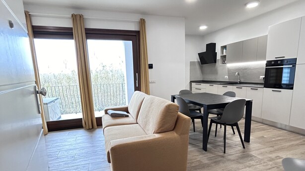 Una sala moderna con un sofá color beige, una mesa de comedor con sillas, una cocina con armarios blancos, y un balcón con cortinas.