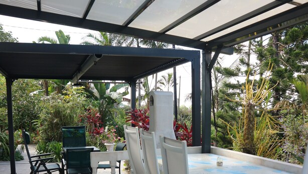 Ein überdachter Patio-Bereich mit Tisch und Stühlen, umgeben von Pflanzen und Bäumen.