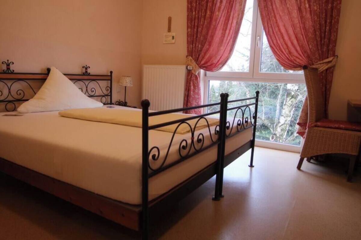 A bedroom with a bed, a window with curtains, a radiator, and a chair.