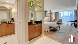 Habitación de hotel con una cama grande, zona de estar con vistas, kitchenette y baño con espejo amplio y tocador.