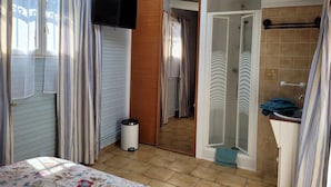 Een kleine kamer met een bed, een aan de muur bevestigde televisie, een douche en een kleine prullenbak.