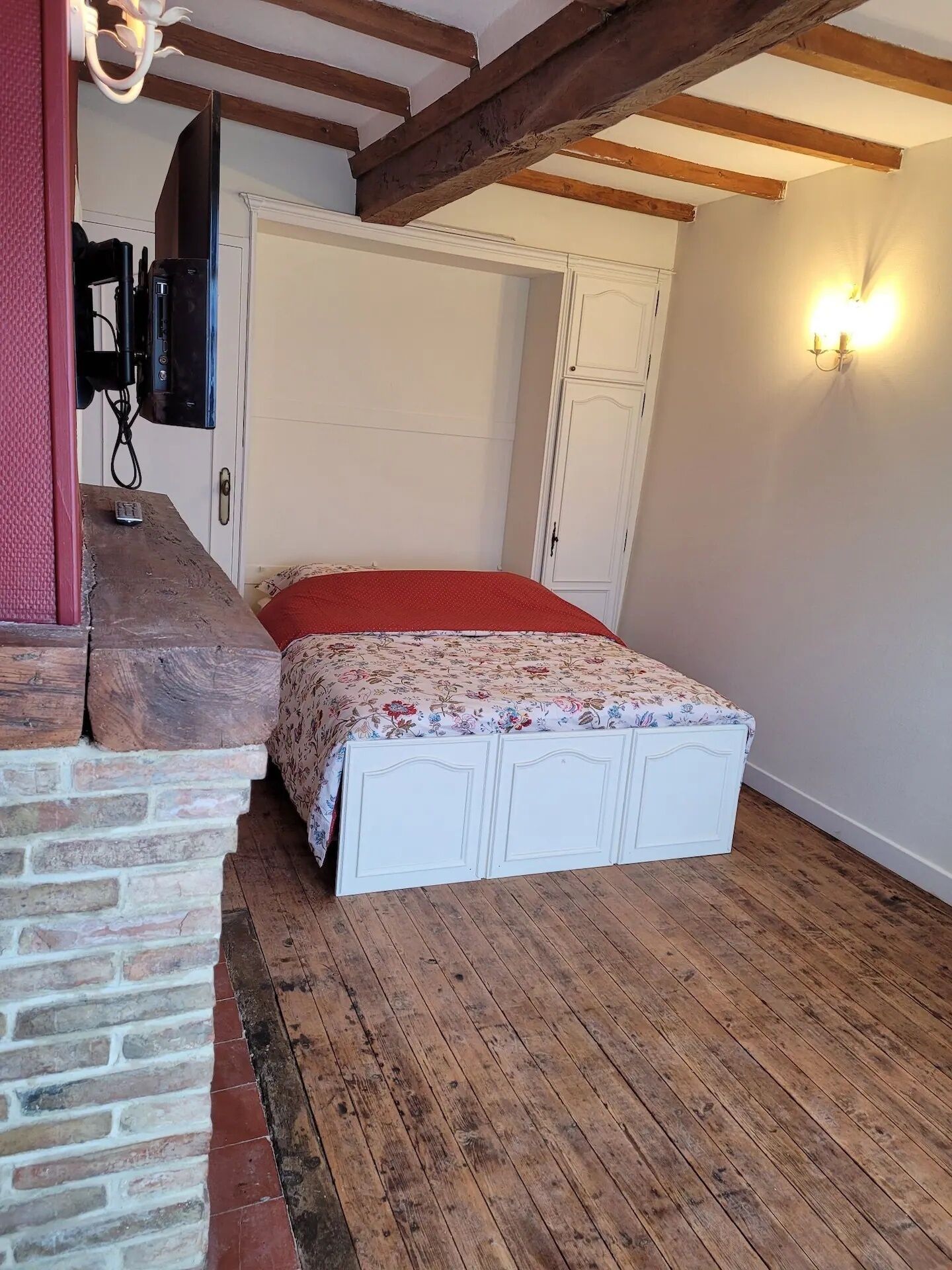 Un dormitorio con cama, suelo de madera, pared de ladrillo, armario blanco y una lámpara fijada a la pared.