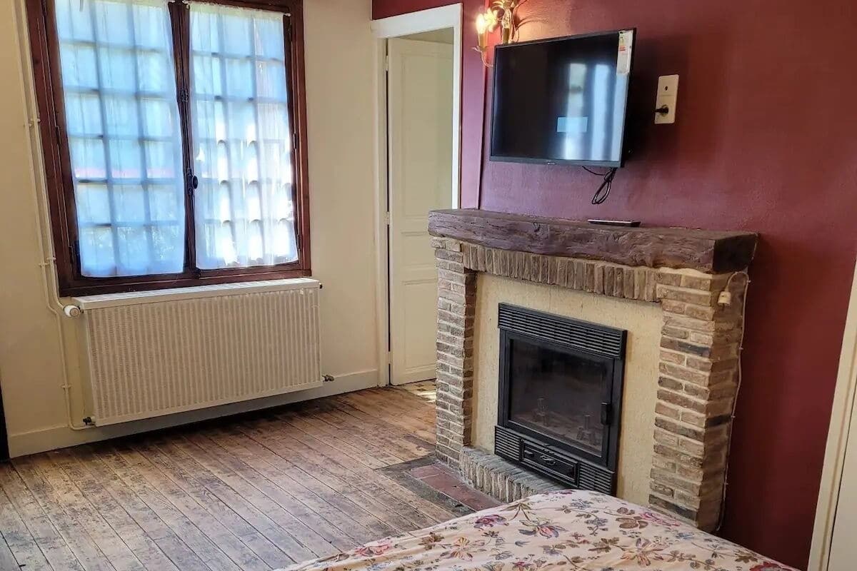 Ein Schlafzimmer mit Kamin, einem Bett mit floralem Überwurf, einem Fenster mit Jalousien und einem Fernseher, der über dem Kamin angebracht ist.