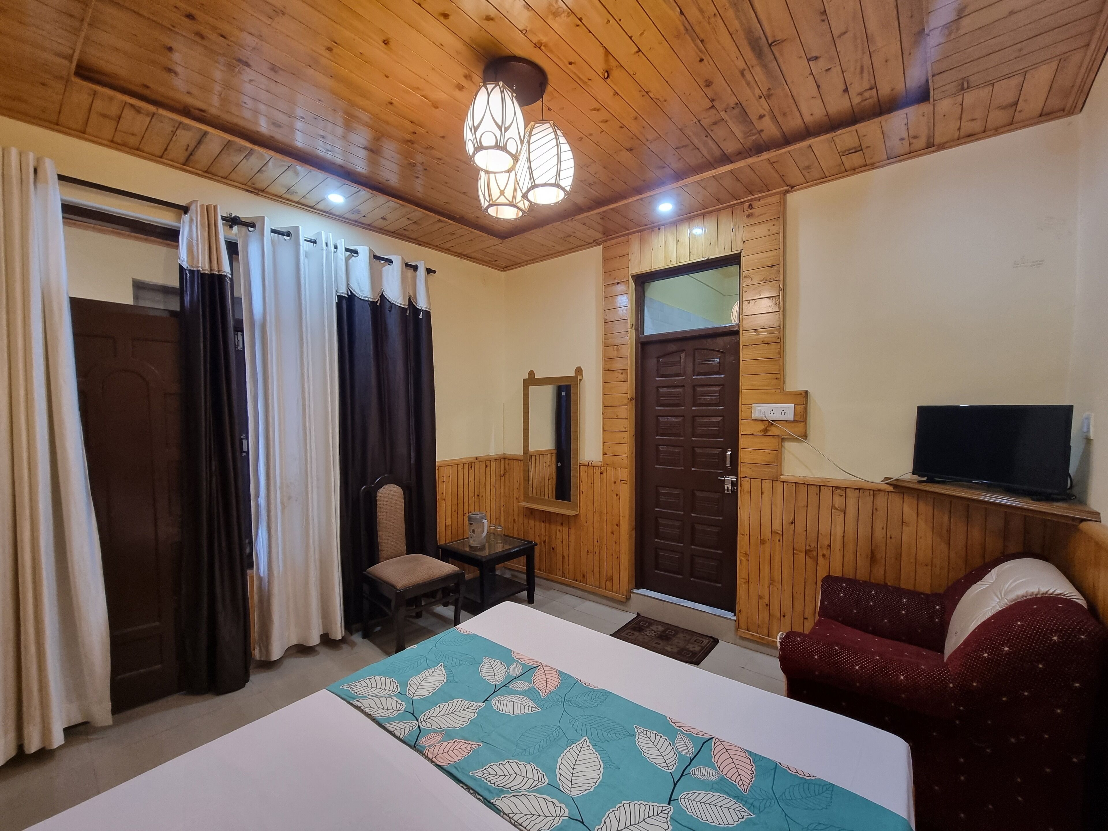 A room with wooden paneling, a bed with a leaf-patterned bedspread, a chair, a small table with a kettle, a television on a shelf, and a door with a mirror above it.