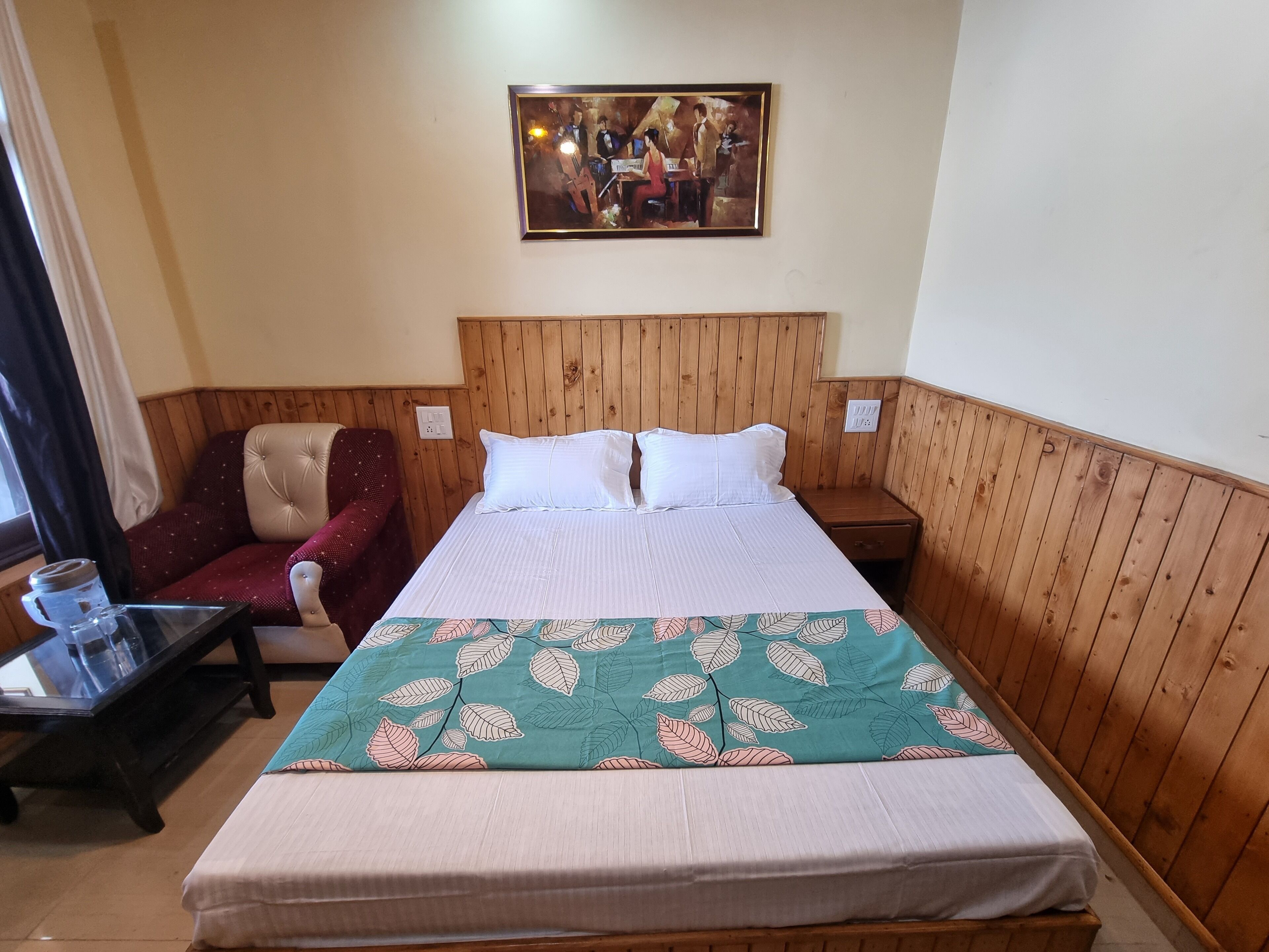 Ein Einzelbett mit Bettdecke im Blattmuster, ein hölzernes Kopfteil, ein Nachttisch, ein Stuhl und ein Bild an der Wand.