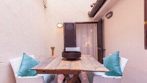 A wooden table with a bowl, surrounded by wicker chairs with teal cushions, in a narrow outdoor space with a door and wall-mounted light fixture. - Campo Viola 10 people (Rome)