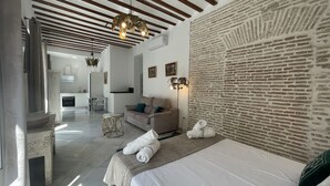Une chambre d’hôtel moderne avec un mur d’accent en briques, un canapé, un lit avec des serviettes, un coin repas et une cuisine en arrière-plan.