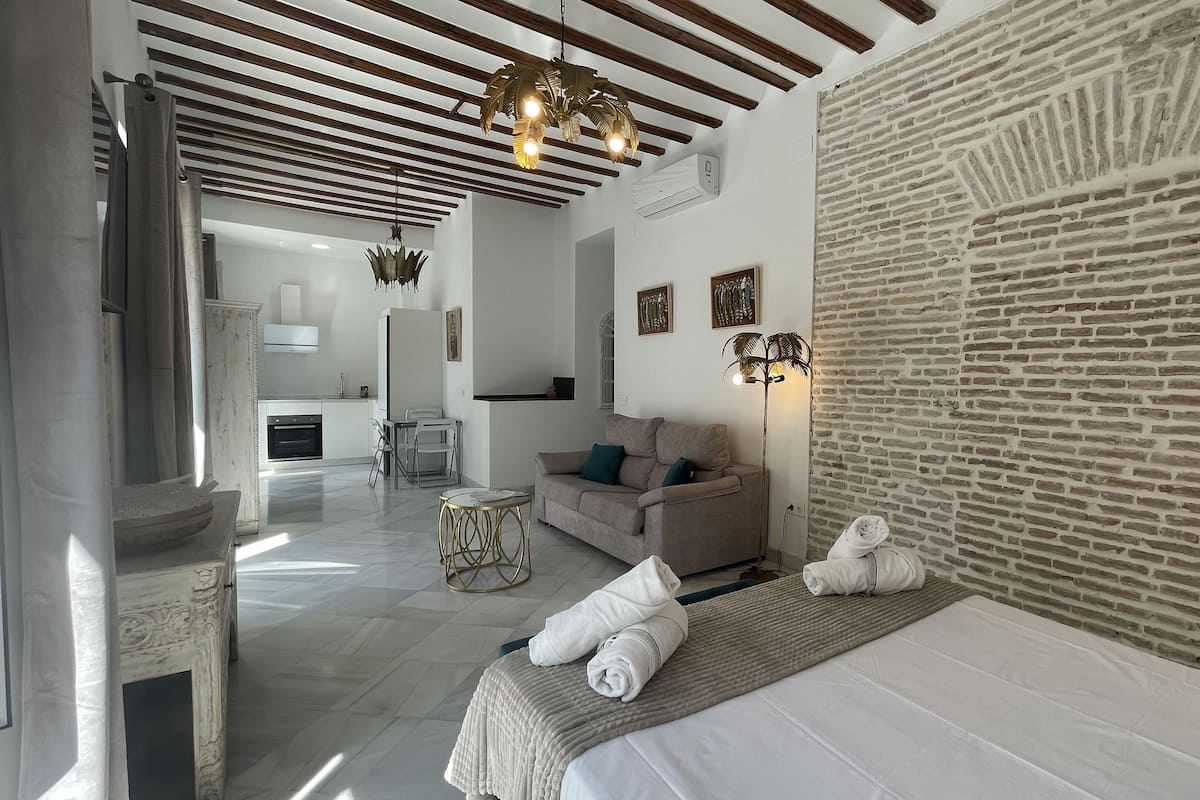 A modern hotel room with a brick accent wall, a sofa, a bed with towels, a dining area, and a kitchen in the background.