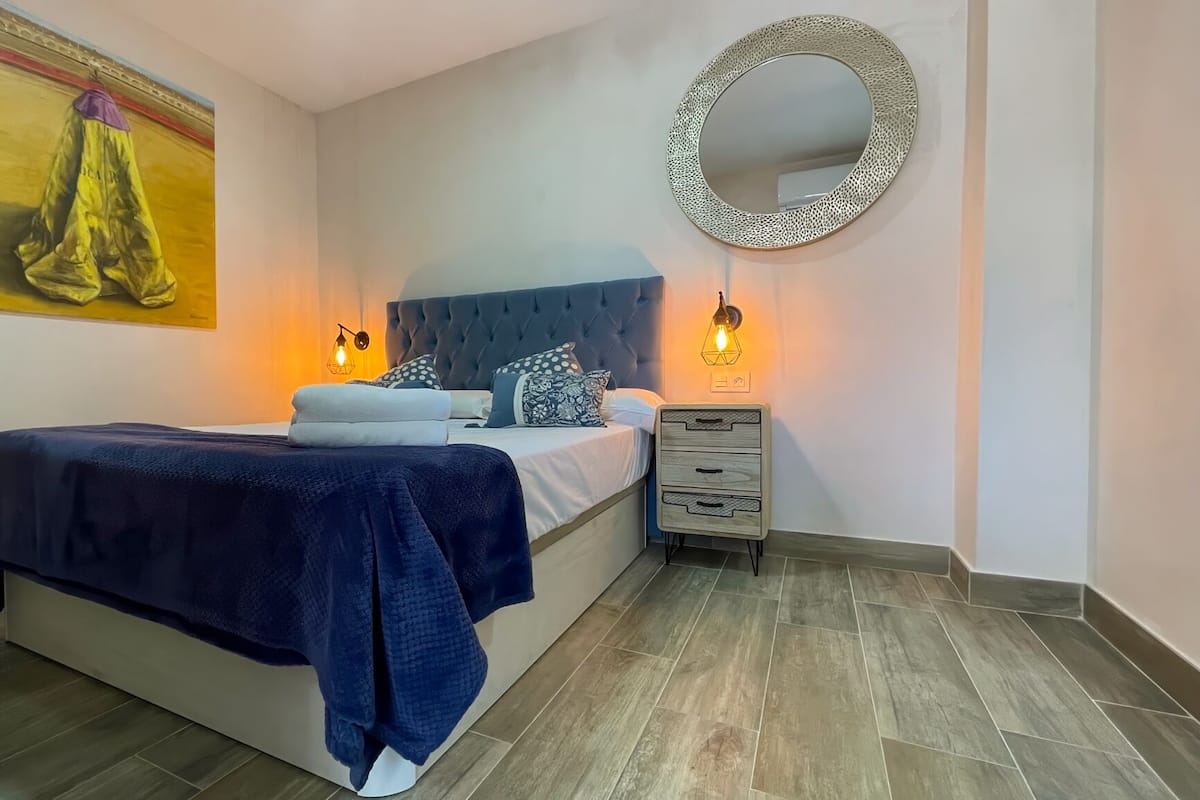 A bedroom with a bed, bedside tables, a wall-mounted mirror, and a painting.