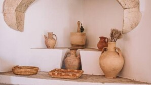 A rustic interior with a stone archway, shelves holding pottery, and a wooden shelf with a jug. - Melody House in Roustika (Rethymno)