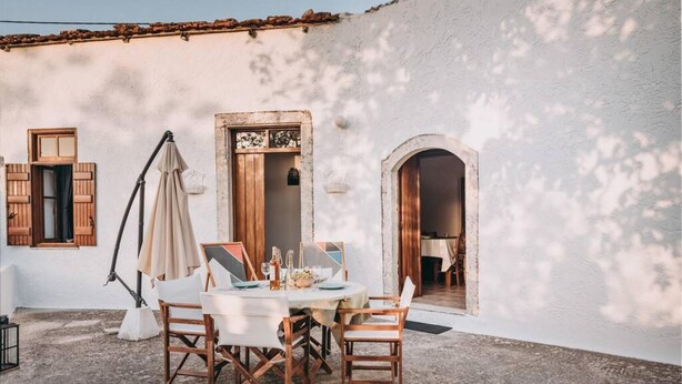 A rustic outdoor dining area with a round table and wooden chairs, set against a white building with wooden shutters and an arched doorway. - Melody House in Roustika (Rethymno)