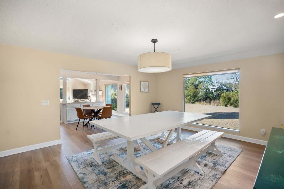 Un comedor amplio con una gran mesa y bancos, una cocina al fondo, y vistas a un jardín a través de ventanas grandes.