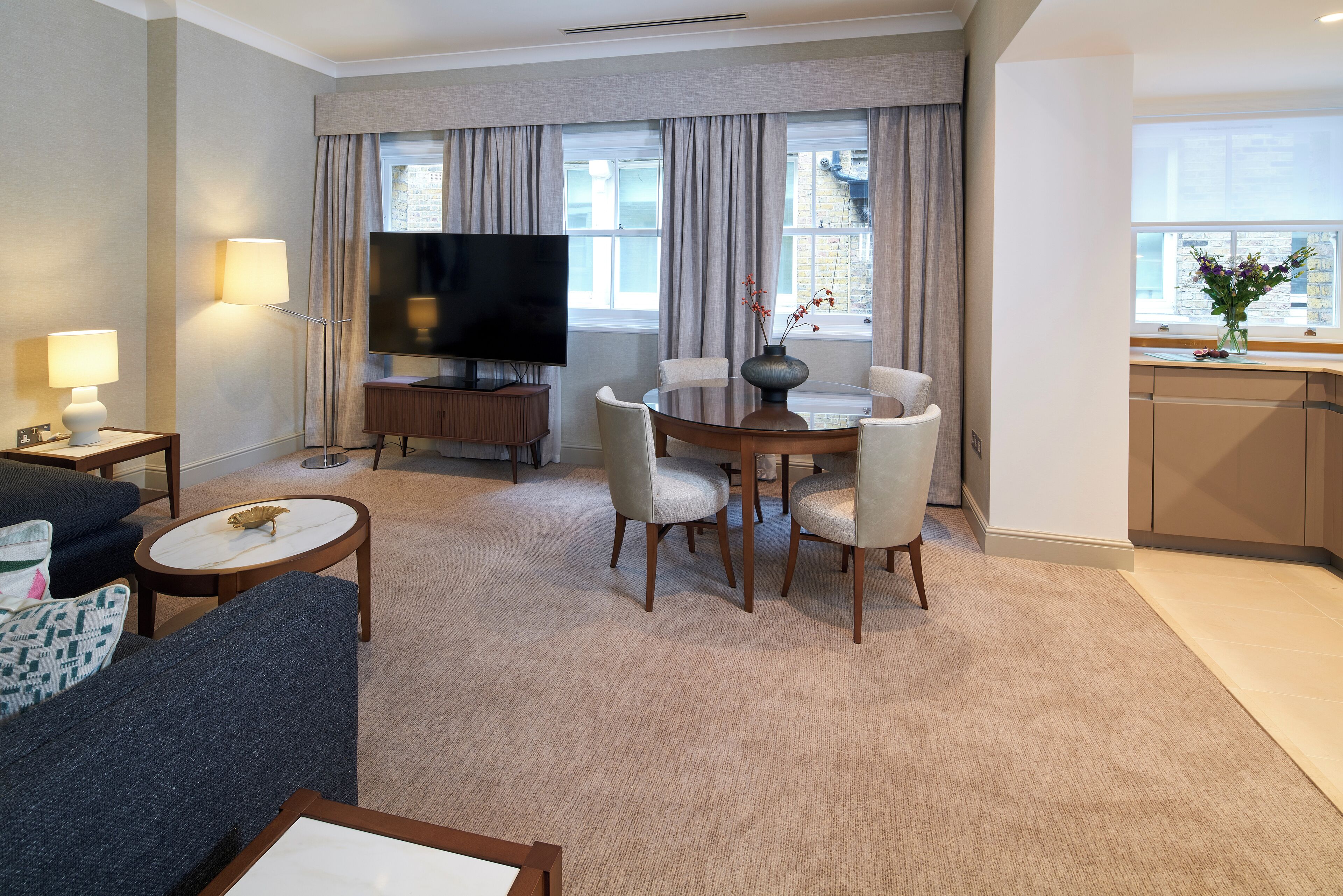A modern hotel room with a flat-screen TV, a dining table with chairs, a sofa, a small round table, and a kitchen area with a vase of flowers.