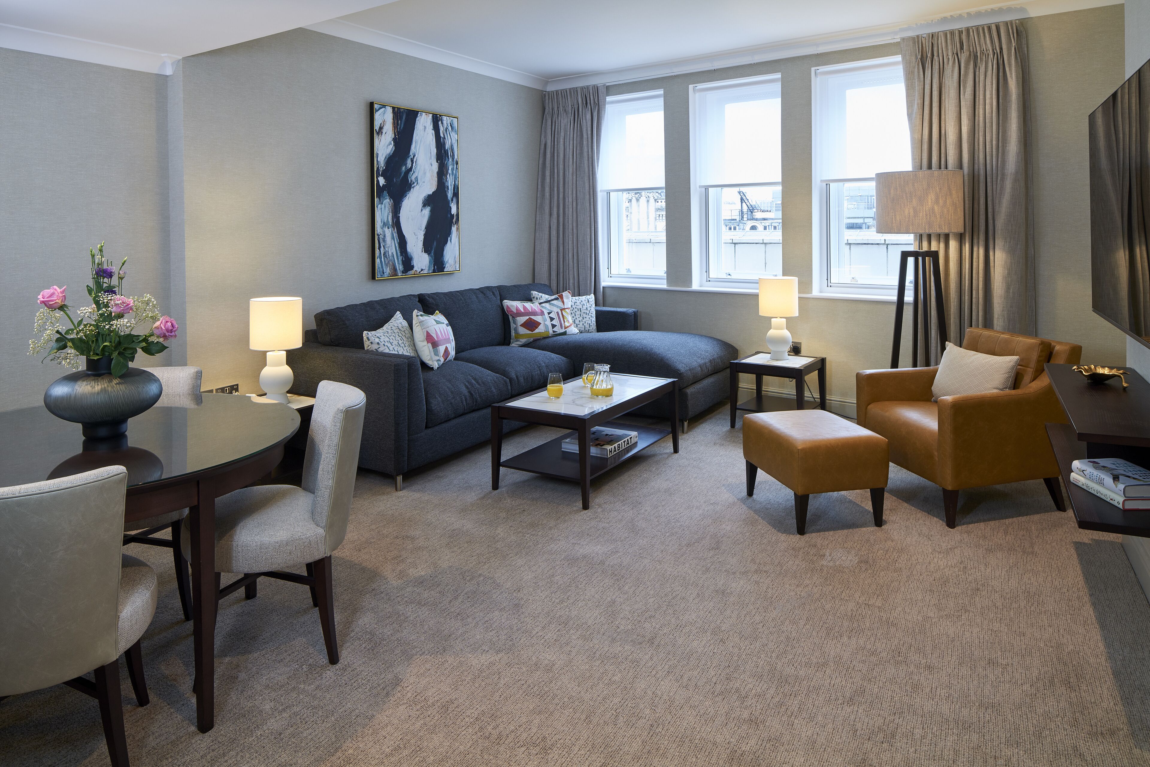 Un salon moderne avec un canapé gris, deux fauteuils, une table basse et une table à manger avec des chaises. Un tableau orne le mur, une lampe sur pied est posée au sol, et de grandes fenêtres offrent une vue sur la ville.