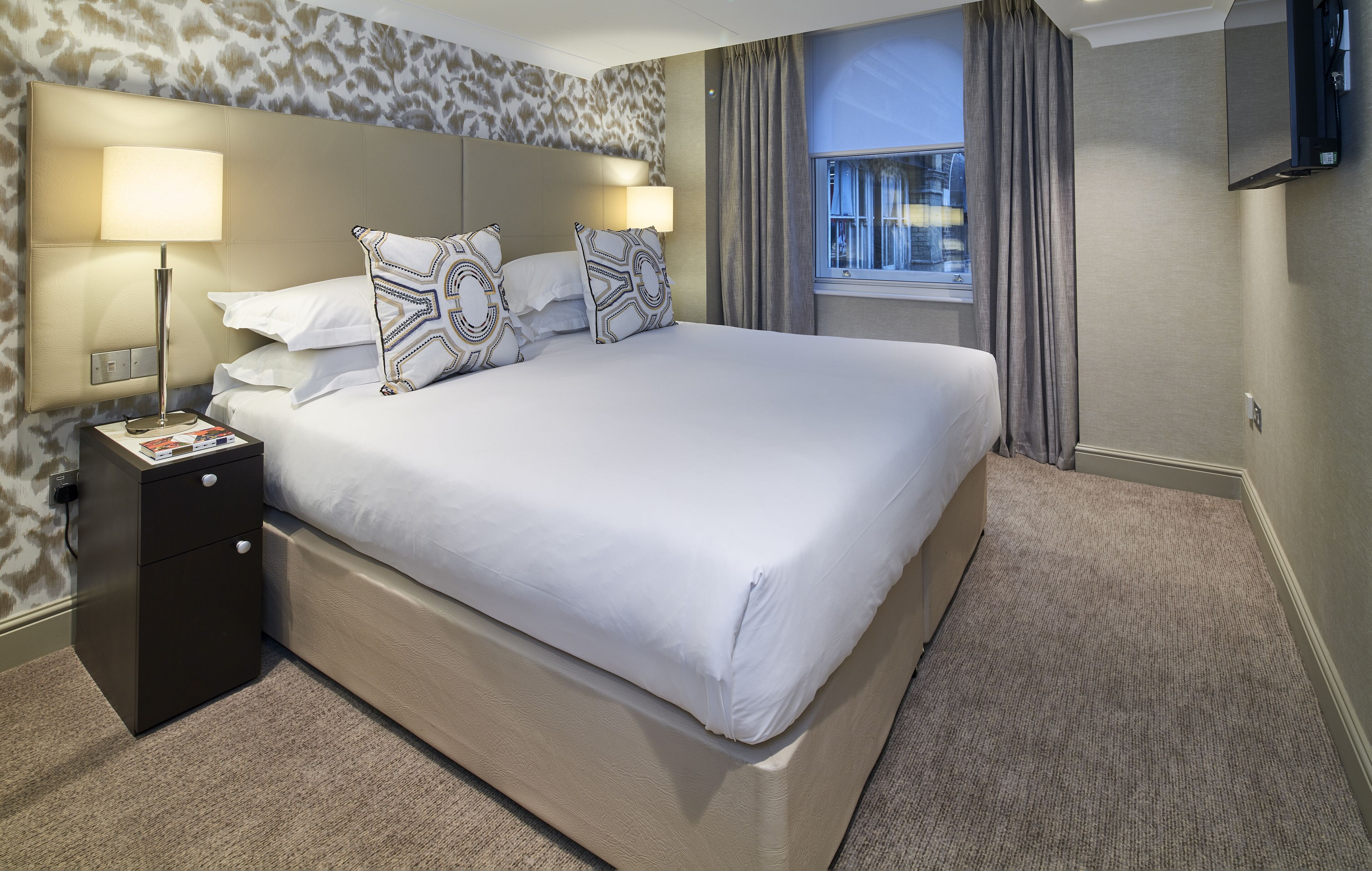 A hotel room with a large bed, a nightstand with a lamp, a TV mounted on the wall, and a window with curtains.