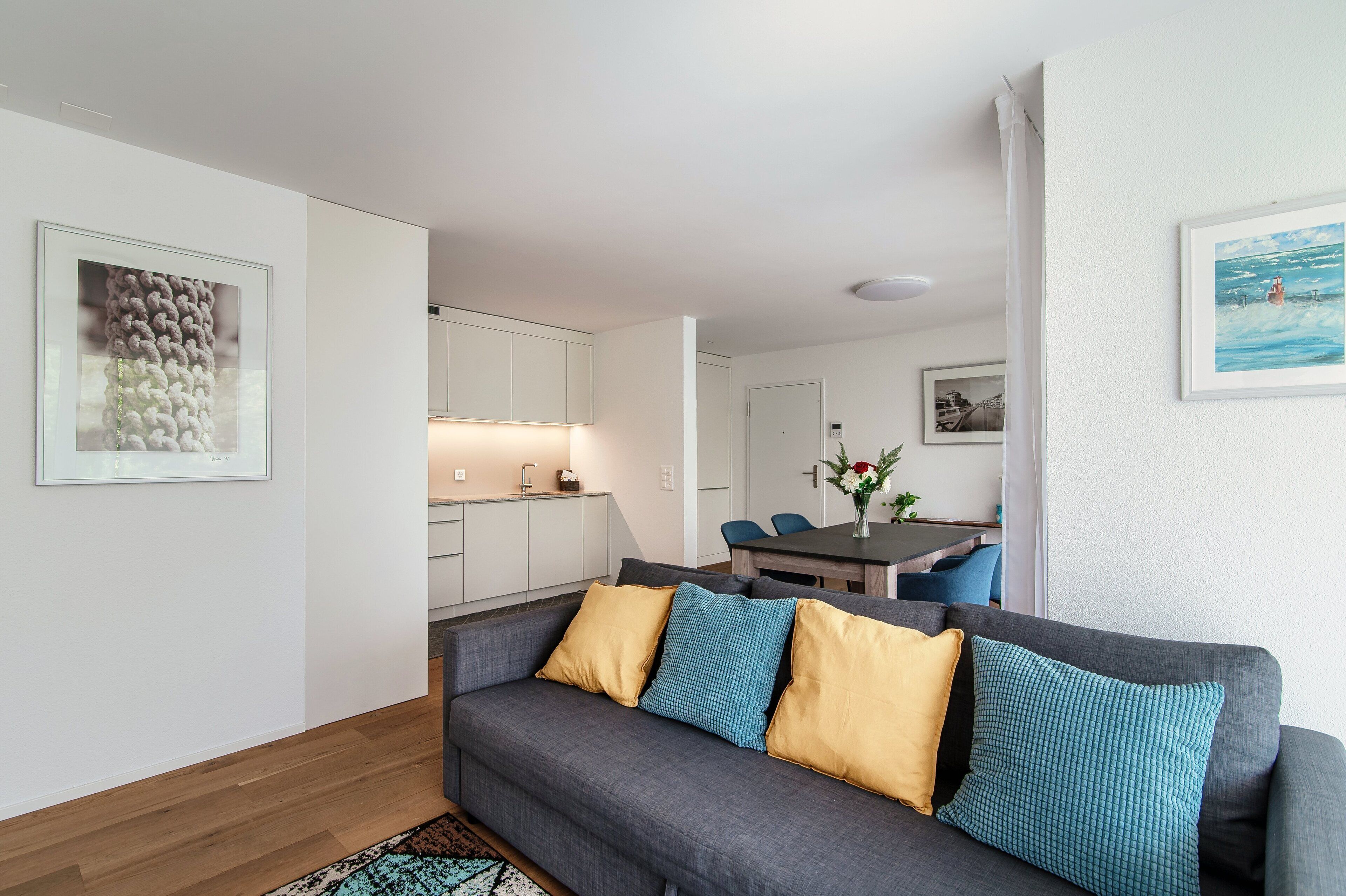 A modern living room with a grey sofa, yellow and blue cushions, a wooden floor, a dining area with a table and chairs, and a kitchen in the background.