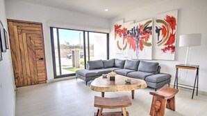 Una sala moderna con un sofá seccional gris, una mesa de centro de madera y dos taburetes de madera. Hay cuadros abstractos en la pared y una lámpara en una mesita auxiliar.