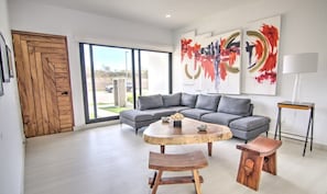 A modern living room with a gray sectional sofa, a wooden coffee table, and two wooden stools. There are abstract paintings on the wall and a lamp on a side table.