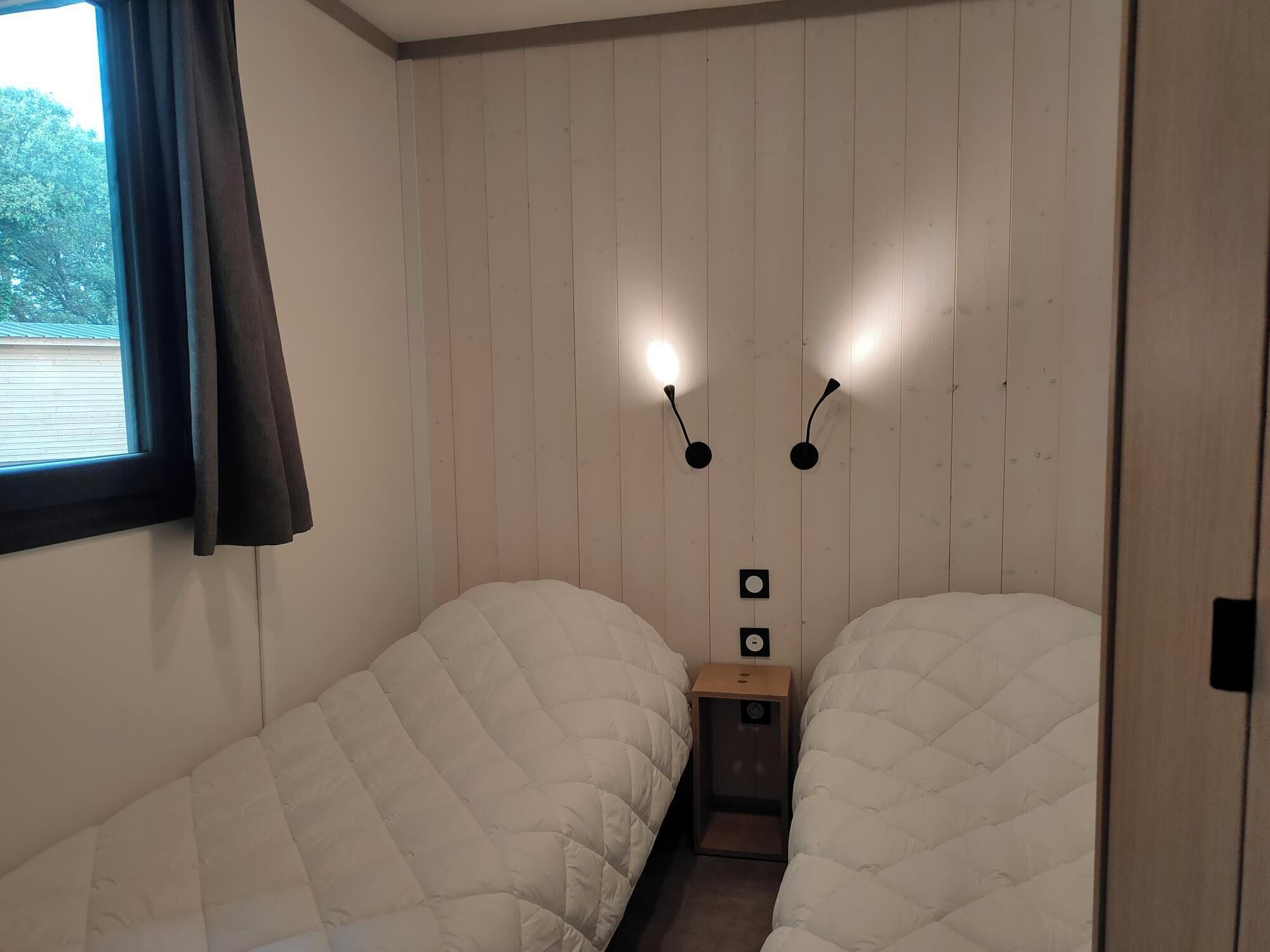 A room with two beds, a window with curtains, and wall-mounted lighting.