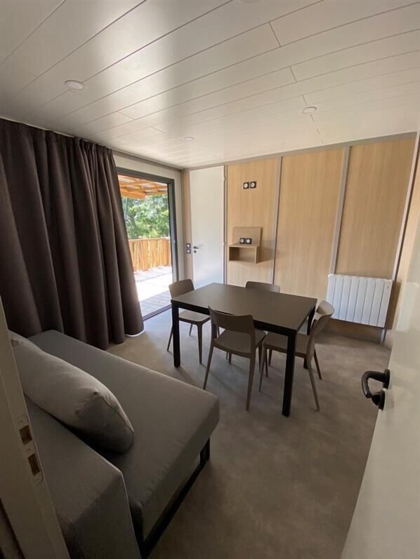 A modern living room with a sofa, a dining table with chairs, a sliding door leading to an outdoor area, and a built-in shelf with two speakers. - VVF Les Sableaux à Noirmoutier (Noirmoutier-en-l'Ile)