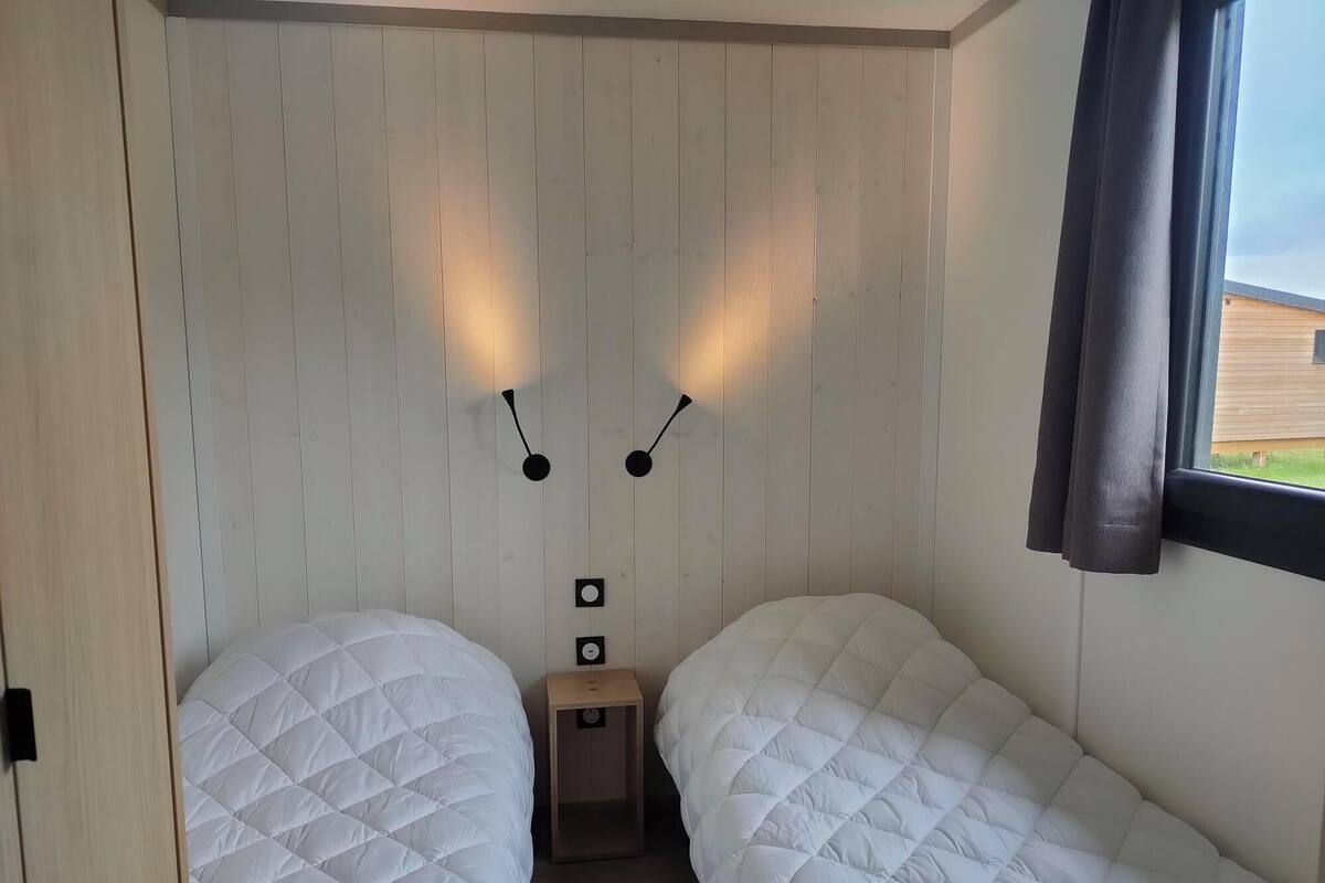 A room with two beds, wooden panel walls, a small bedside table, and a window with curtains.