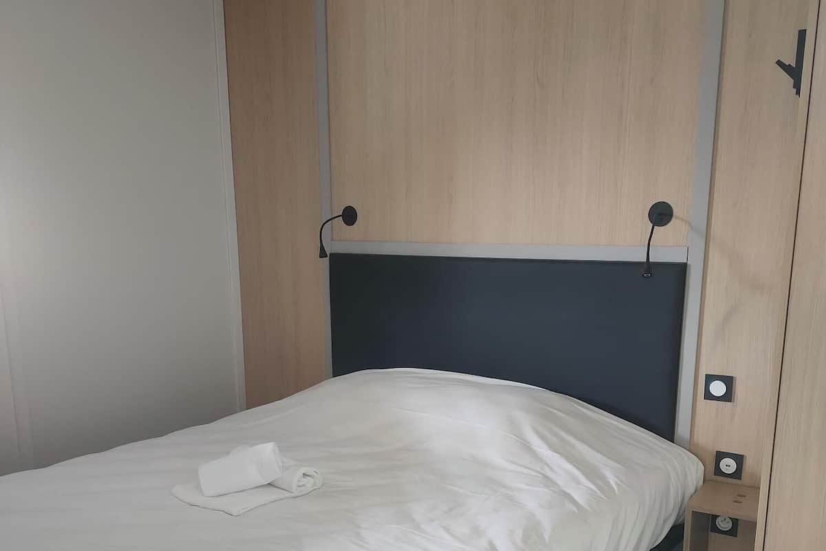 A single bed with a white mattress and a neatly folded towel, a wooden wardrobe with a dark headboard, and a small bedside table with a lamp.