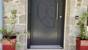 Une porte de couleur sombre ornée d’un motif circulaire, flanquée de deux jardinières rouges contenant des plantes vertes.