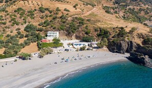Vue aérienne d’une propriété située sur la côte, avec une plage, un bâtiment comprenant plusieurs pièces et un espace de stationnement.