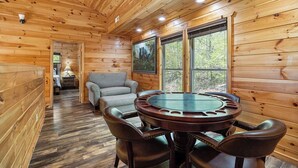A wooden cabin interior with a round dining table, leather chairs, a sofa, and large windows offering a view of the forest. - Upscale 4 Bedroom Amazing View, Hot Tub, Arcade/Theater, No Winding Roads! (Sevierville)