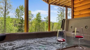 A hot tub with two glasses of red wine, surrounded by wooden beams and large windows overlooking a forest. - Upscale 4 Bedroom Amazing View, Hot Tub, Arcade/Theater, No Winding Roads! (Sevierville)