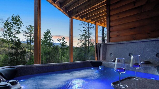 A hot tub with two glasses of red wine, surrounded by wooden beams and large windows overlooking a forested landscape. - Upscale 4 Bedroom Amazing View, Hot Tub, Arcade/Theater, No Winding Roads! (Sevierville)