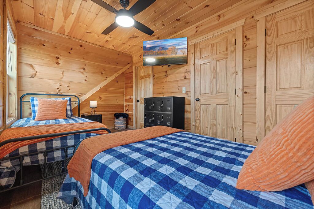A cozy cabin bedroom with two beds, wooden walls, a ceiling fan, a television, and a small cabinet.