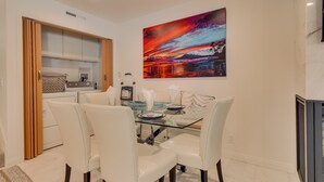 A dining area with a glass table and white chairs, a painting of a sunset on the wall, and a kitchen with wooden cabinets and a washing machine.