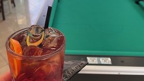 A hand holding a glass with a drink, a pool table, and a view of a pool area with lounge chairs. - Hotel Apogeo  (Rimini)