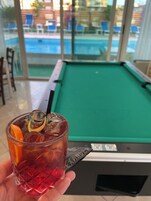 Una mano sosteniendo un vaso con una bebida, una mesa de billar y vistas a un área de piscina con sillones de descanso.