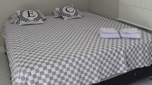 A single bed with a checkered bedspread, two pillows with monogrammed letters, a folded towel, and a wall-mounted air conditioning unit.