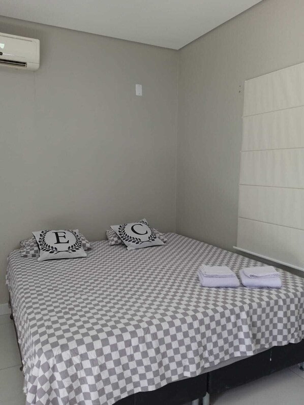 A single bed with a checkered bedspread, two pillows with monogrammed letters, a folded towel, and a wall-mounted air conditioning unit.